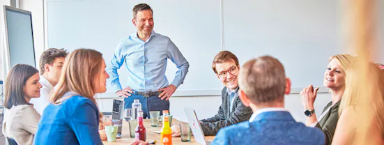 Christopher Hasperg steht lächelnd vor einer Tafel, während KollegInnen sitzend zuhören und interagieren.