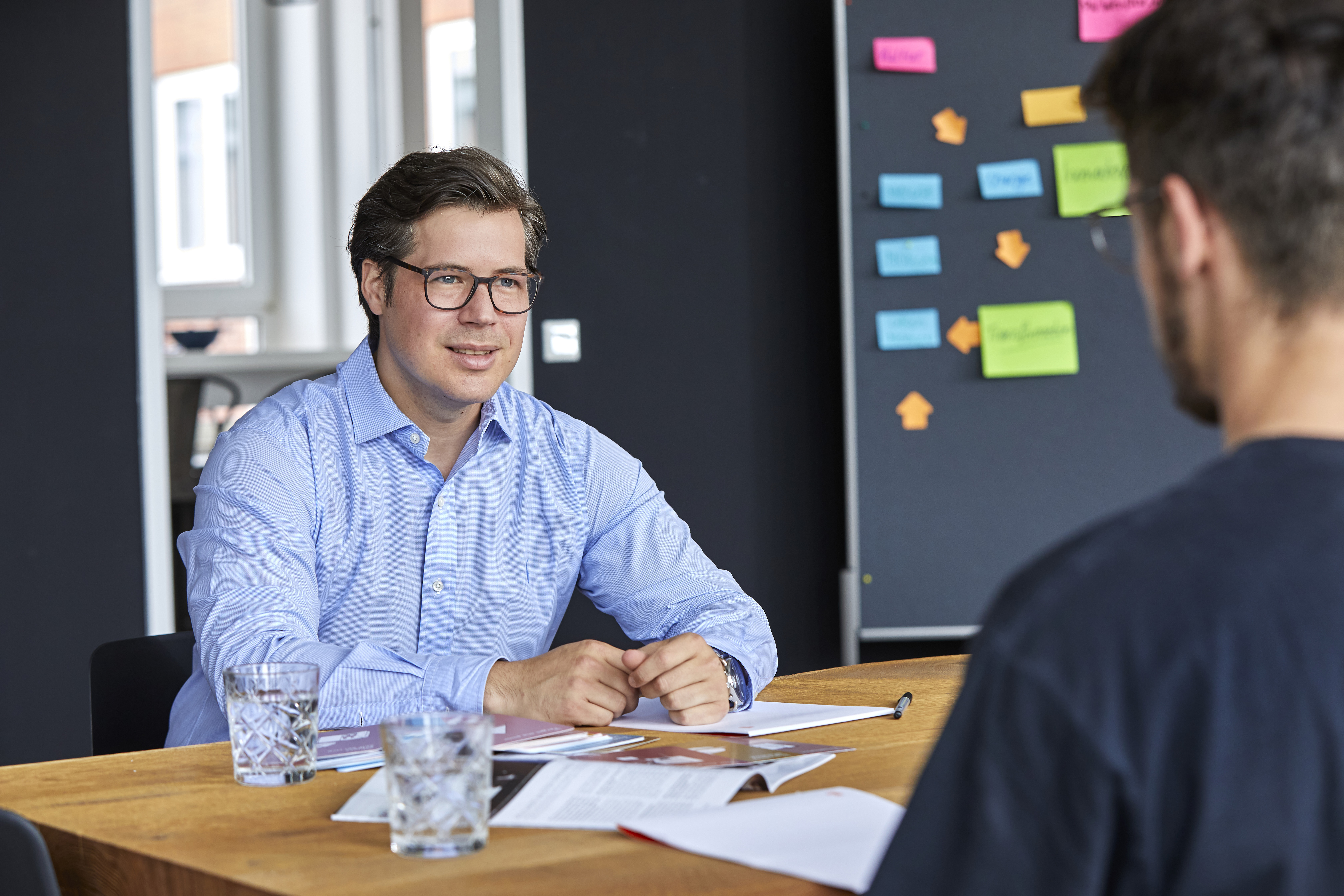 Ein Mitarbeiter der Allfoye sitzt an einem Tisch und unterhält sich mit einem Mann. Der Mitarbeiter hat dunkle Haare, trägt eine Brille und ein blaues Hemd.