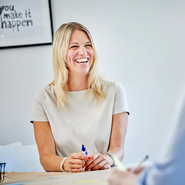 Eine Mitarbeiterin sitzt an einem Tisch und lacht gemeinsam mit ihrem Gesprächspartner. Beide wirken entspannt und haben offensichtlich Spaß an der Unterhaltung