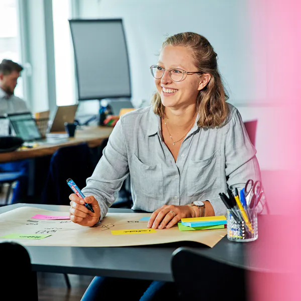 Eine Mitarbeiterin arbeitet an einem Plakat, hält einen Stift in der Hand und lächelt breit. Sie wirkt kreativ und begeistert bei ihrer Aufgabe.