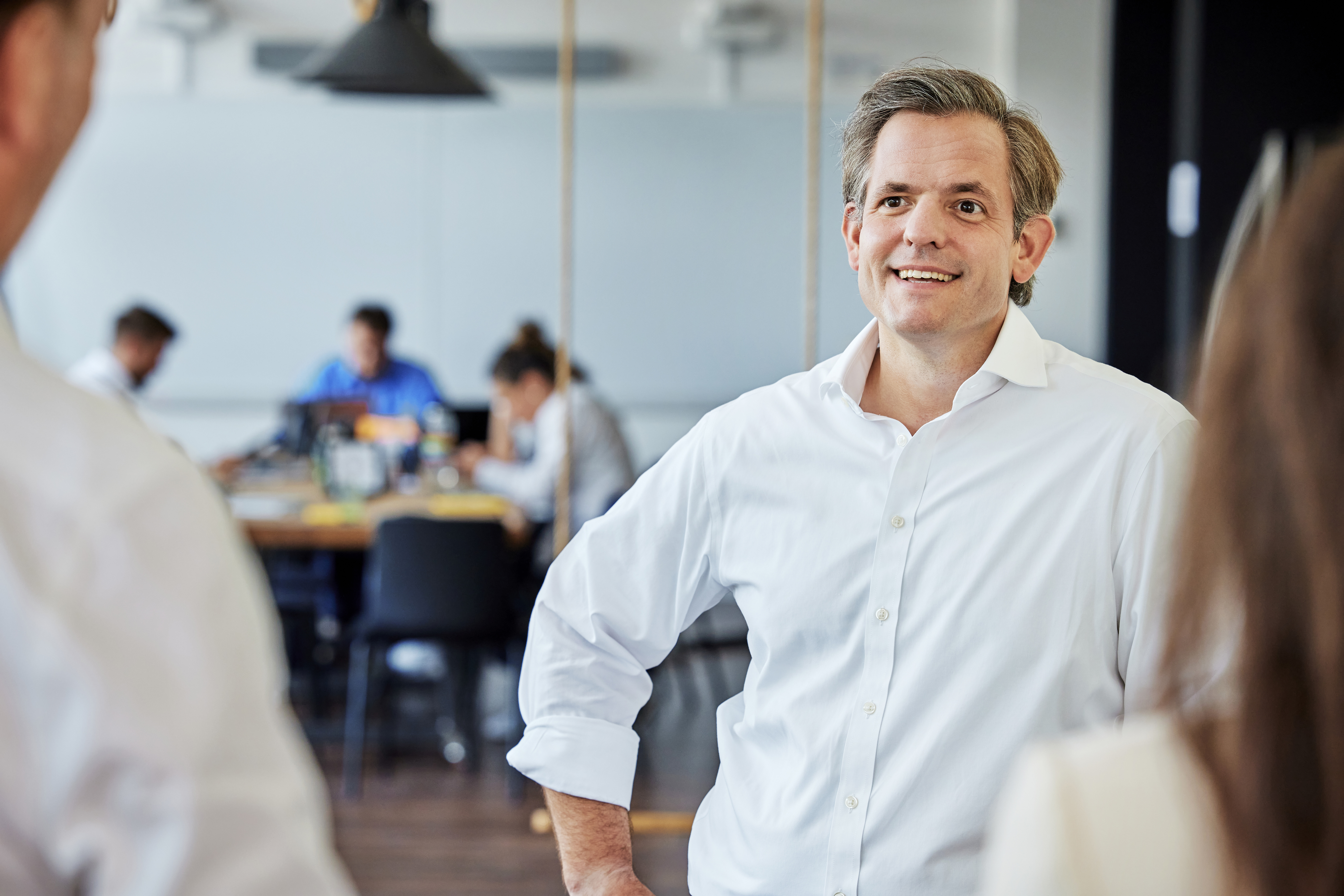 Der CEO unterhält sich lachend mit einem Mitarbeiter, beide stehen miteinander. Der CEO steht im Fokus des Bildes und wirkt entspannt und in einem freundlichen Gespräch mit dem Mitarbeiter.