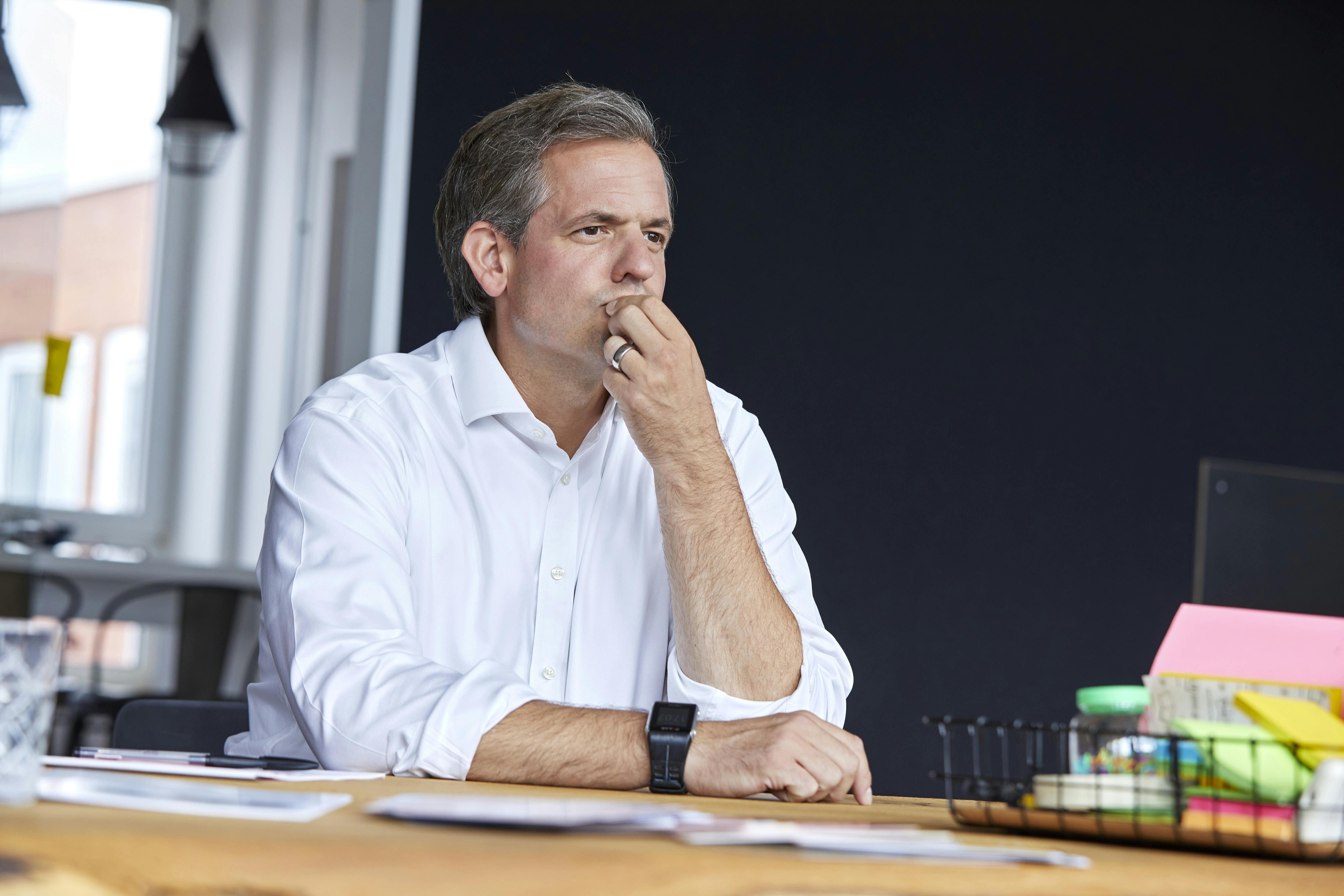 Der CEO sitzt an einem Tisch und schaut nachdenklich in die Ferne. Seine Körpersprache wirkt ruhig und fokussiert, als ob er über eine wichtige Entscheidung nachdenkt.