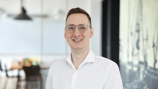 Portrait eines Mitarbeiters mit braunen Haaren, weißem Hemd und einer Brille vor hellem Hintergrund in einem kreativ gestalteten Raum.