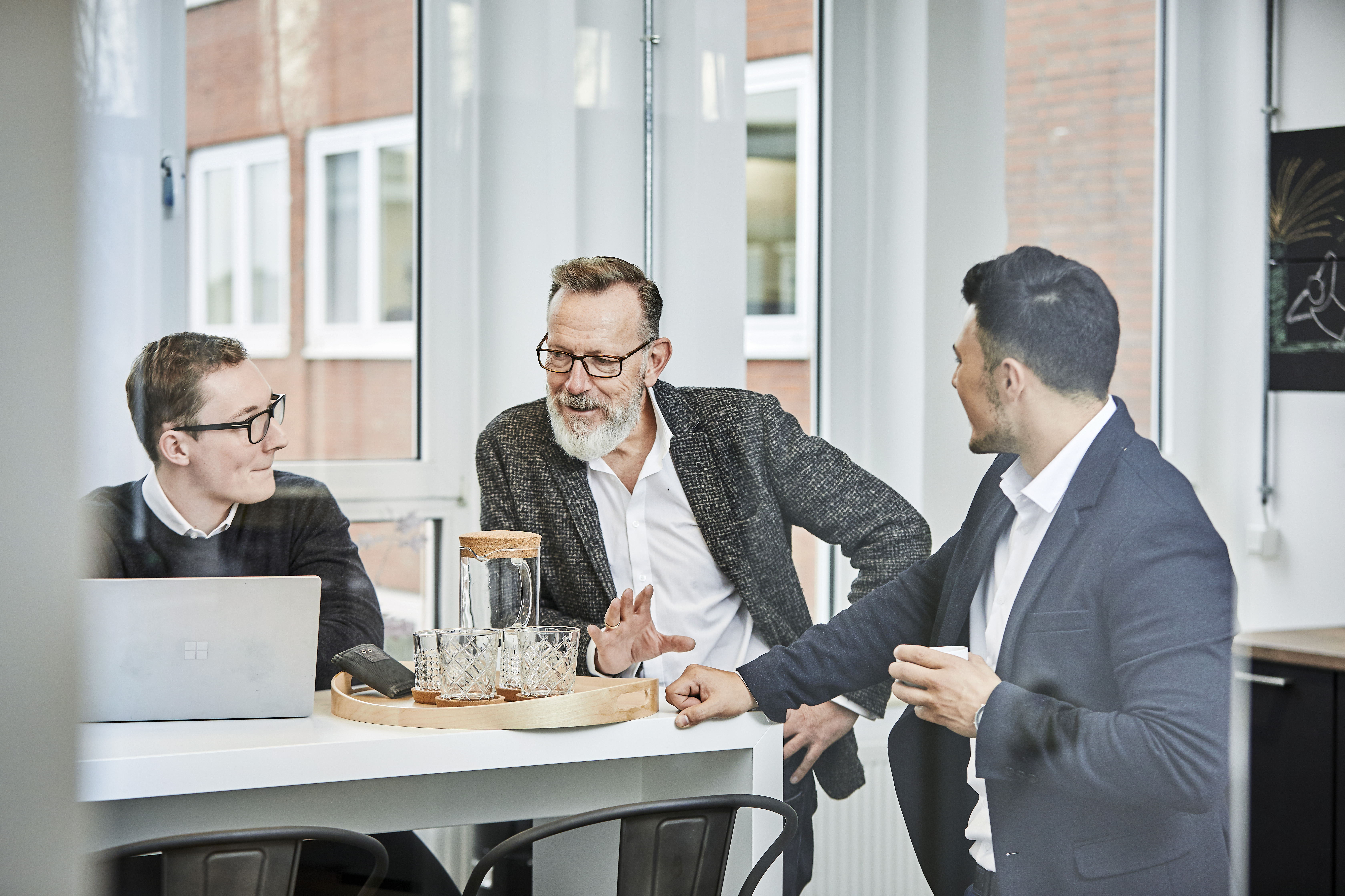 Drei Mitarbeiter stehen an einem Stehtisch in einer Küche und besprechen konzentriert etwas auf einem Laptop. Die Atmosphäre wirkt professionell und kollegial, während sie gemeinsam an einer Aufgabe arbeiten.