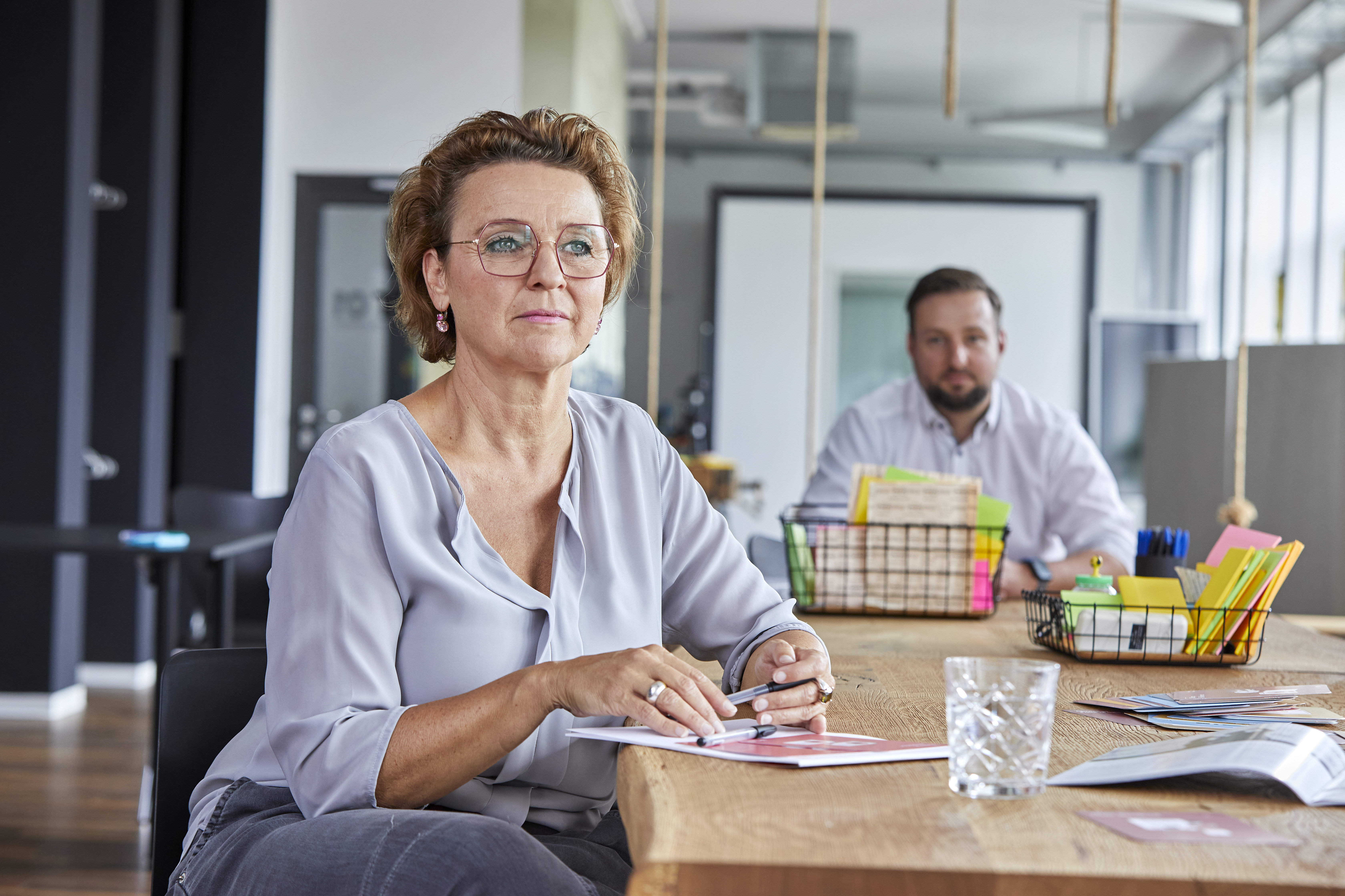 Eine Mitarbeiterin sitzt an einem Tisch und wirkt konzentriert, während sie zuhört. Im Hintergrund ist ein männlicher Kollege zu erkennen, dessen Präsenz dezent bleibt, sodass der Fokus auf der Frau liegt.