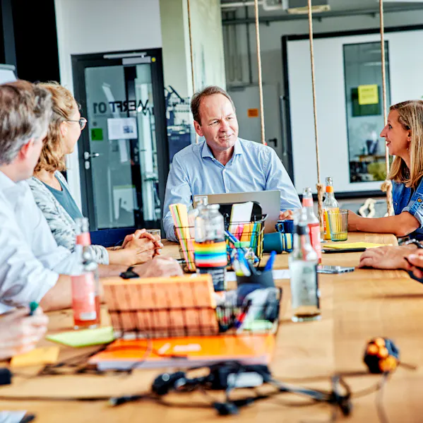 Eine Gruppe von Personen ist in einem Raum um einen großen Tisch versammelt. Papiere und Laptops liegen verstreut herum.