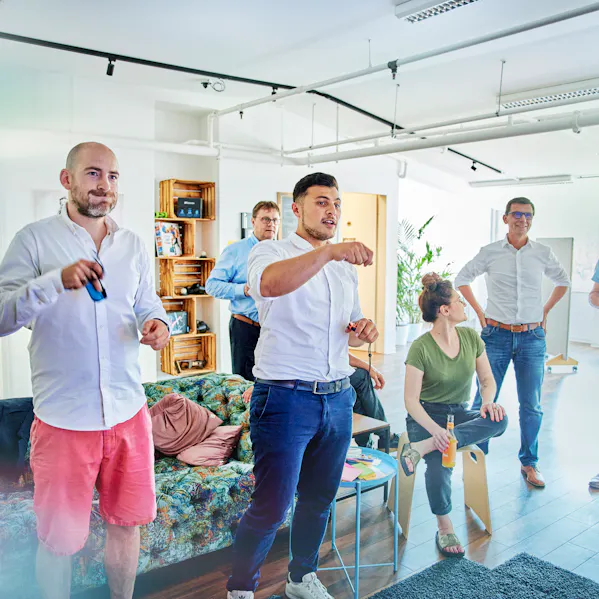 Einige Mitarbeiter stehen an einer Konsole und spielen gemeinsam, sichtlich begeistert und vertieft. Im Hintergrund sitzen andere Kollegen auf einer Couch, unterhalten sich und haben ebenfalls Spaß, was eine lockere und fröhliche Atmosphäre vermittelt.