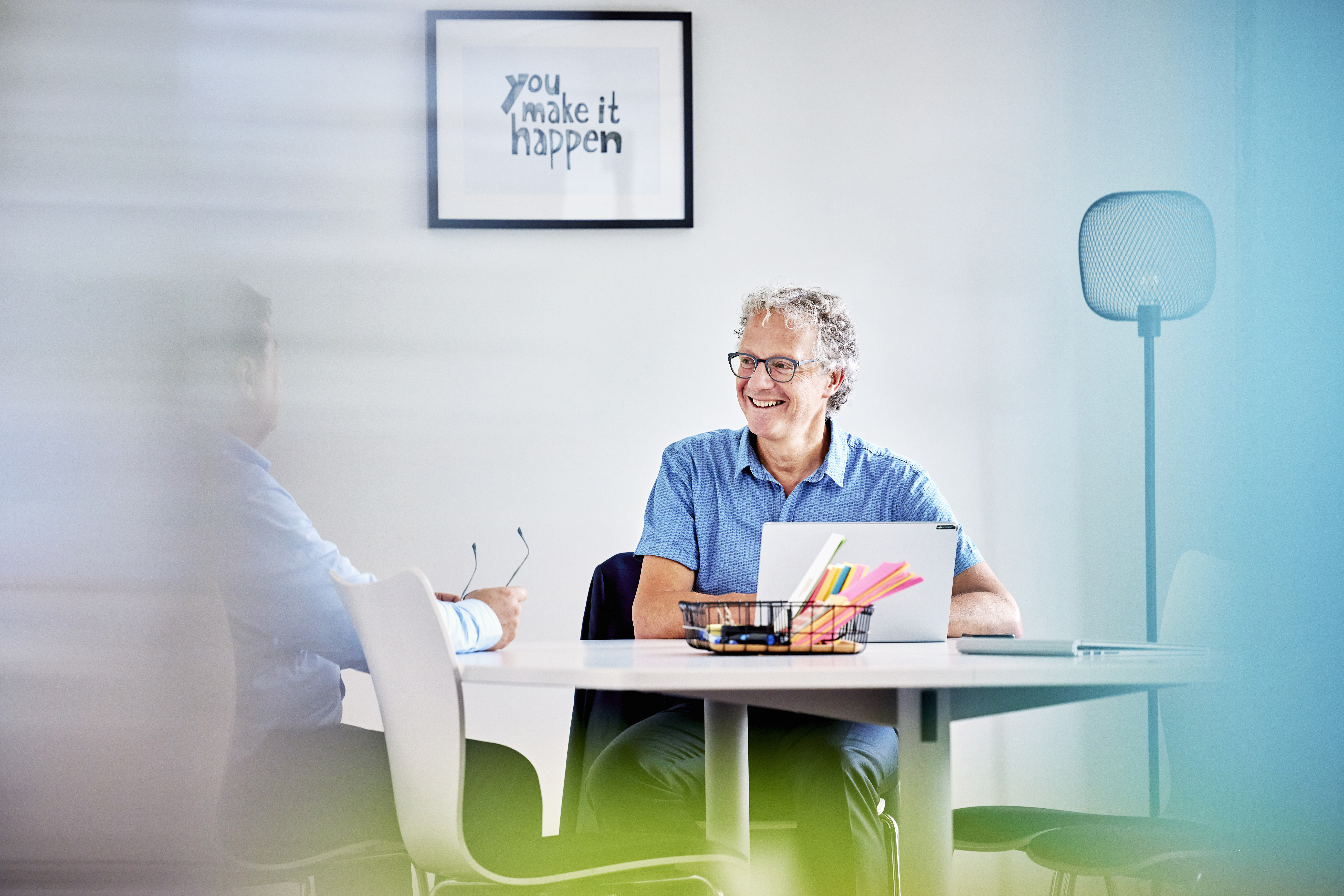 Ein Mann sitzt am Tisch im Fokus des Bildes und unterhält sich mit Mitarbeitenden in lockerer Atmosphäre.
