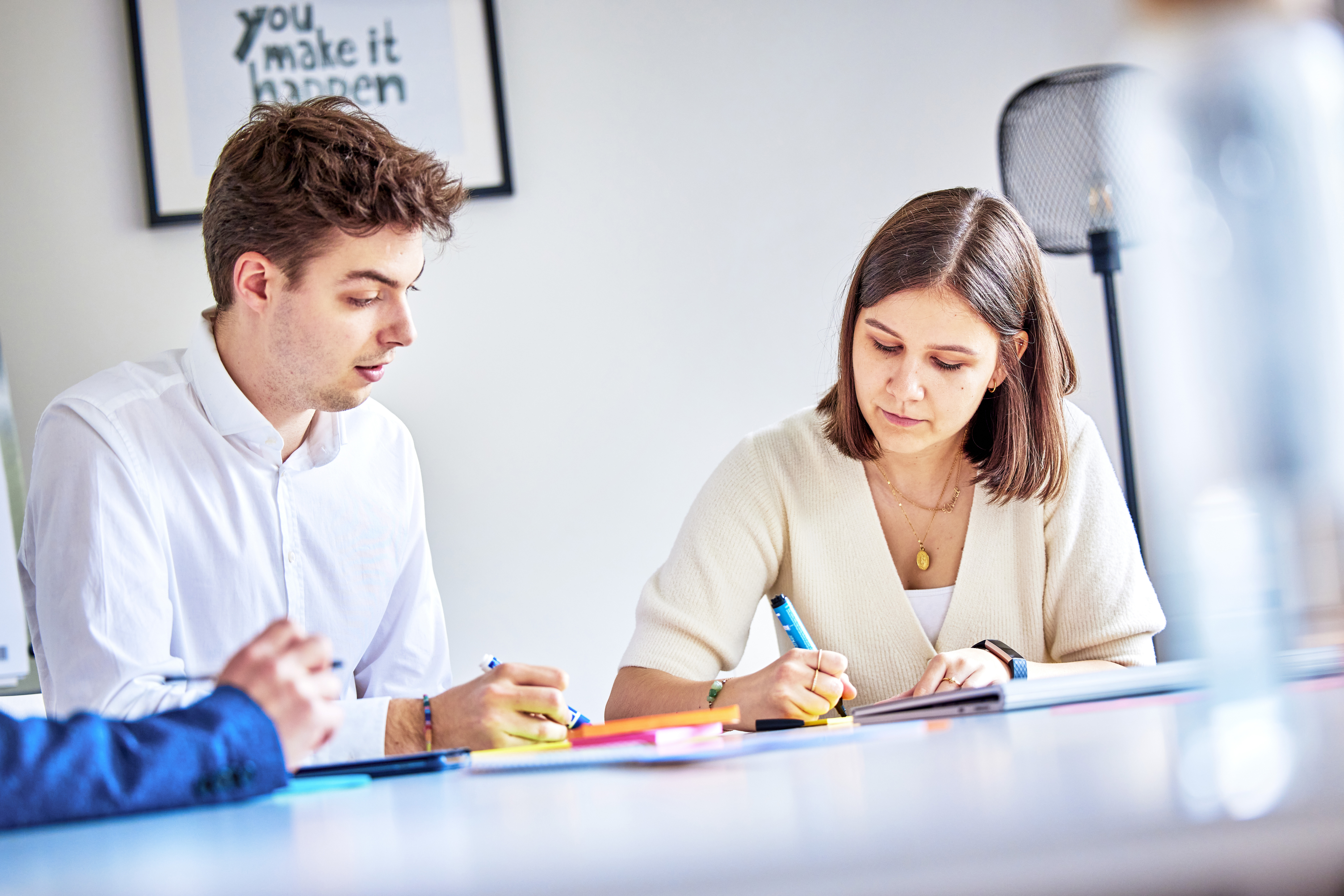 Zwei junge Personen sitzen an einem Tisch, beide tragen helle Kleidung. Sie sind in eine gemeinsame Arbeitssitzung vertieft.