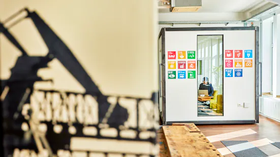 Büroraum mit Nachhaltigkeits-Symbolen der UN SDGs an der Wand und einer Person beim Arbeiten am Laptop.