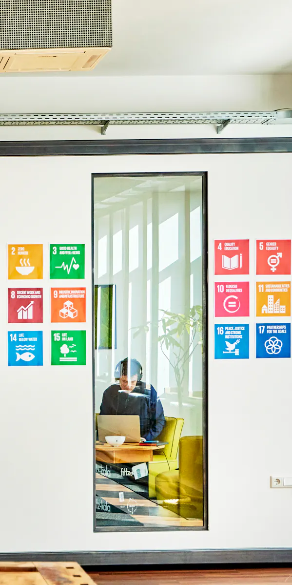 Moderne Büroszene mit einer Person, die in einem kleinen, verglasten Raum an einem Laptop arbeitet. An der Wand hinter ihr hängen bunte Symbole der 17 UN-Nachhaltigkeitsziele. Im Vordergrund unscharf ein industrielles Wandmotiv, das einen Kontrast zur hellen, offenen Arbeitsumgebung bildet.