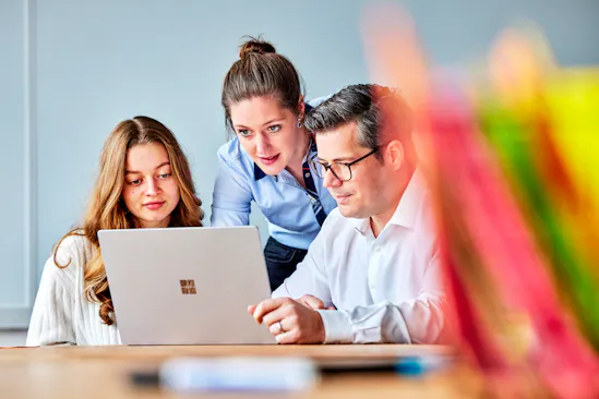 Drei Personen schauen auf einen Laptop und arbeiten dort virtuelle mit anderen Personen zusammen