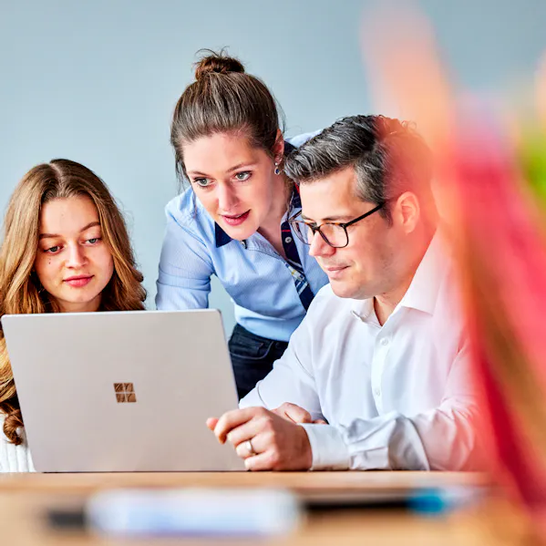 Drei Personen schauen auf einen Laptop und arbeiten dort virtuelle mit anderen Personen zusammen