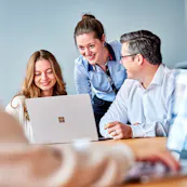 Drei lächelnde Mitarbeitende betrachten konzentriert einen Laptop. Zwei sitzen nebeneinander, die mittlere Person steht und beugt sich interessiert über den Bildschirm.