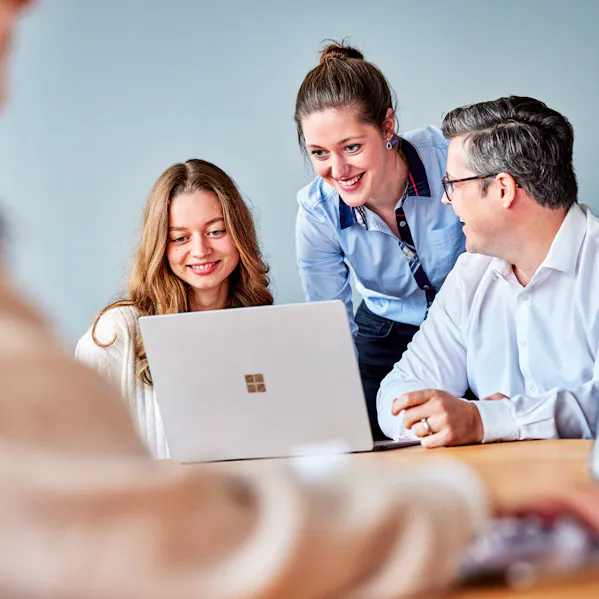 Ein kleines Team sitzt am Tisch, die mittlere Person beugt sich interessiert über den Laptop, während alle fröhlich in den Bildschirm blicken.