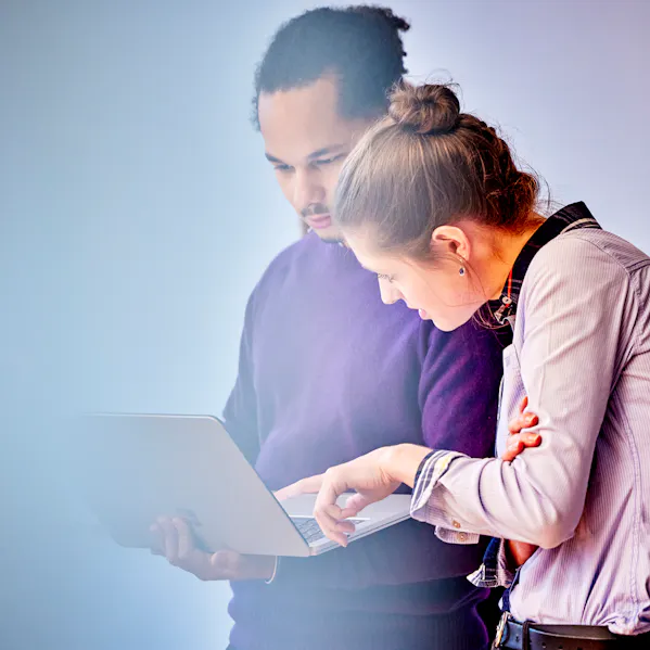 Ein Mann und eine Frau stehen nebeneinander in einem Raum und blicken gemeinsam auf einen geöffneten Laptop. Sie arbeiten konzentriert zusammen.